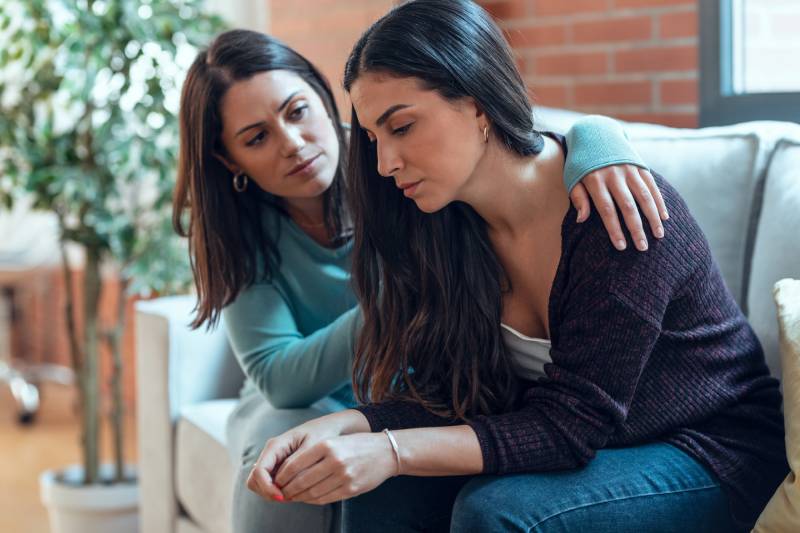 woman comforting her sad friend while sitting on the sofa at home
